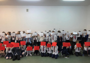 Uczniowie szkoły ubrani na galowo. Stojący uczniowie w rękach trzymają białe kartki, a kucający uczniowie czerwone. Kartki w dłoniach uczniów symbolizują barwy flagi Polski.