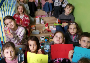 Dzieci siedzą w rzędach na schodach. Na środku, pomiędzy nimi, ustawione są produkty spożywcze, zgodnie z piramidą zdrowego żywienia. Dziewczynki w najwyższym rzędzie trzymają plakat z piramidą. Dzieci siedzące najniżej trzymają kartki z nazwami aktywności fizycznej.