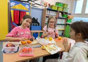 Trzy dziewczynki z klasy 3c siedzą przy ławce, przygotowują szaszłyki owocowe