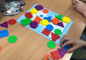 Ręce dzieci, które gają w grę na spostrzegawczość. Dopasowywanie figur geometrycznych po rzucie kostkami (jedna kostka - kształt figury, druga kostka - kolor figury).