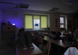 Przyciemniona sala i uczniowie leżący i siedzący przy i na ławkach - relaksacja po lekcjach przy kolorowym świetle lampki.