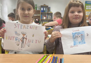 Dwie dziewczynki trzymają w rękach kartki z narysowanymi strażnikami internetu.