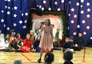 Dziewczynka w różowej sukience śpiewająca przez mikrofon na tle występujących dzieci.