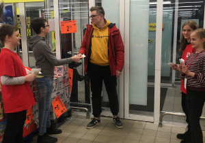 Wolontariusze w wejściu do sklepu Biedronka rozdają ulotki zachęcające do udziału w akcji.