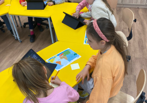 Dzieci parami siedzą przy stolikach i wykonują zadania na tabletach