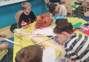 Grupa dzieci siedzi przy stołach i robi latawce z kolorowych kartonów.