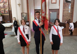 Chłopiec ubrany na galowo trzyma sztandar obok stoją dwie dziewczynki